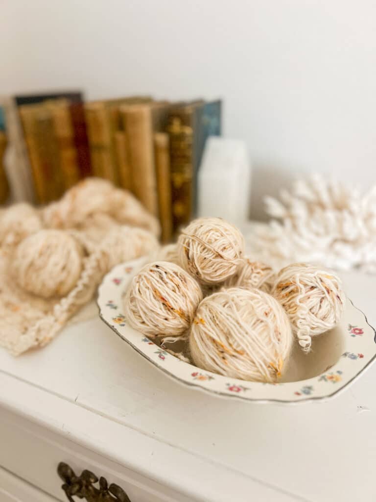 A bowl full of frogged yarn balls sits on top of a white dresser, with a partially frogged sweater and some antique books in the background.