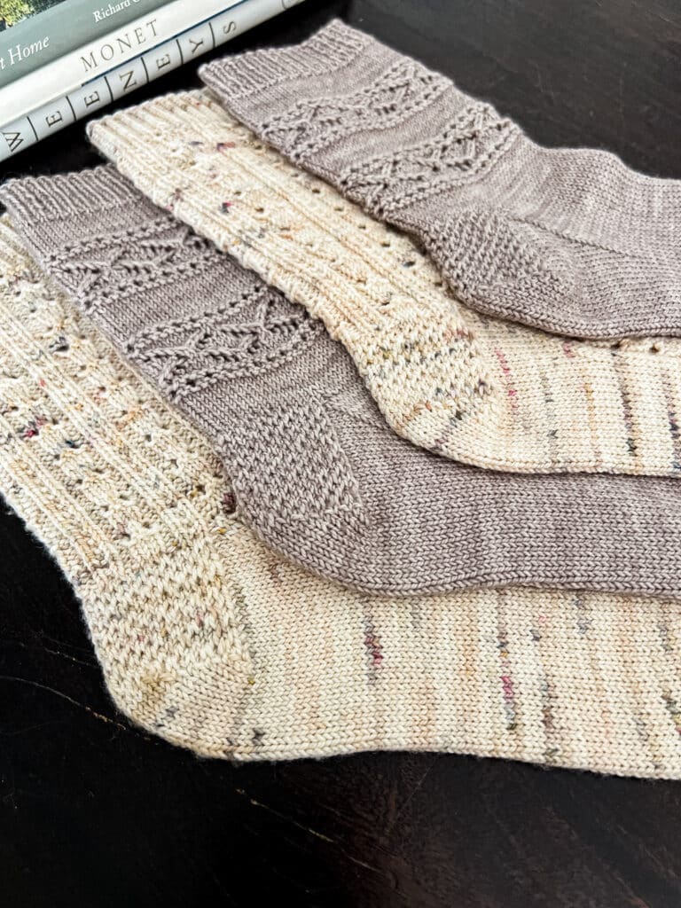 A low-angle photo of four knit socks with Eye of Partridge heels laid out flat on a dark wood table with books in the background. The front heel is the most in-focus.