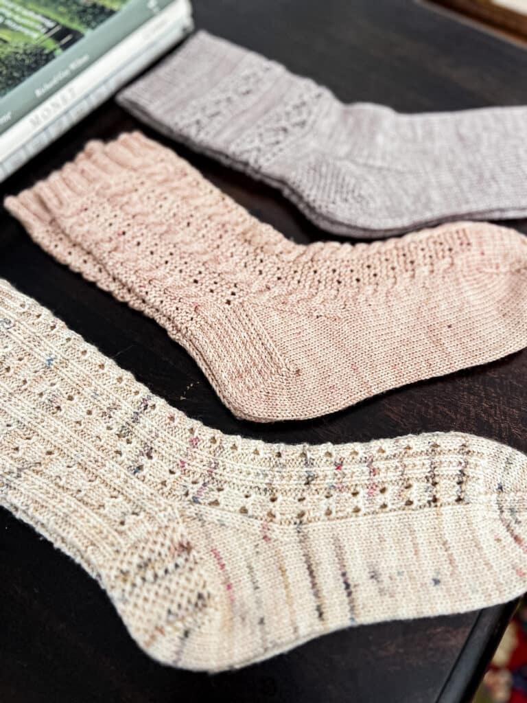 Three pairs of socks are laid out flat on a dark wood table with some books in the background. The front and back pairs have Eye of Partridge heels, while the middle pair has a more traditional heel with stacked slipped stitches.