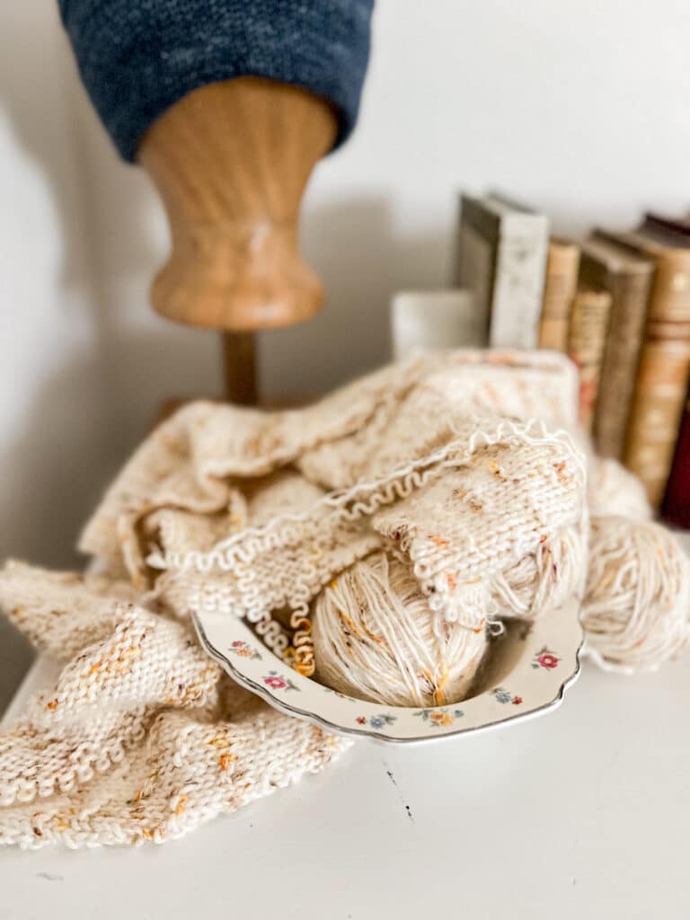 An unfinished, partially frogged sweater sits in a porcelain bowl with little flowers painted on it. In the background, there are antique books and a wooden head form with a knit hat on it.