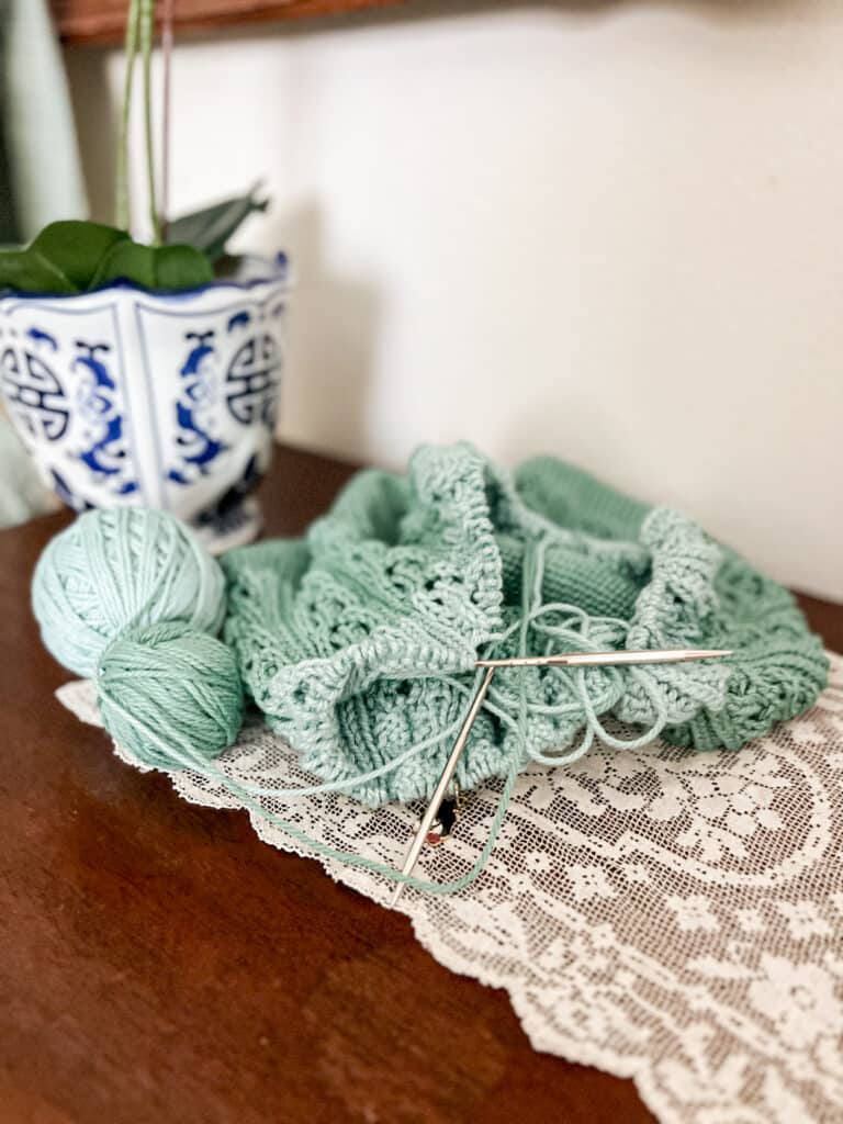 A turquoise and light teal striped knit bag in progress sits on top of a white lace doily on a dark wood tabletop.