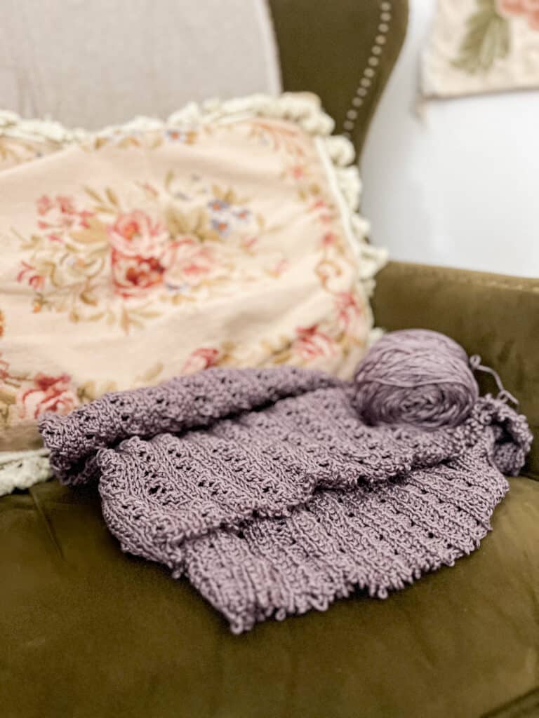A half-knit purple tshirt sits on a dark green wingback chair in front of a floral needlepoint cushion.