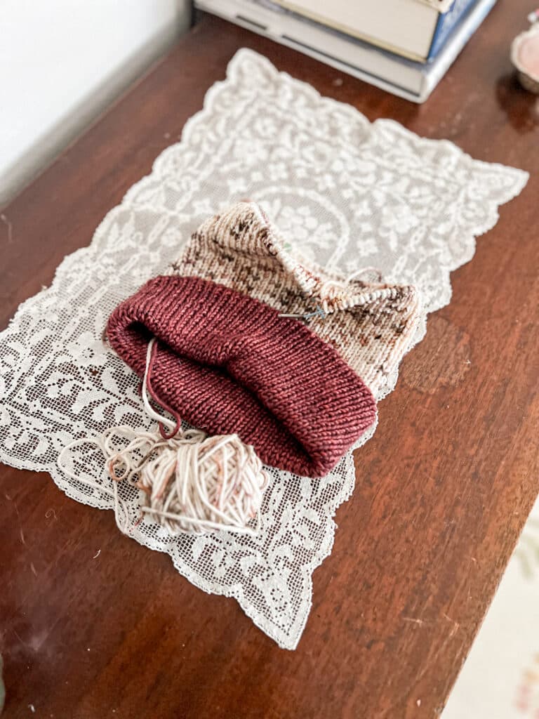 An unfinished 1x1 ribbed had with a burgundy brim and speckled crown sits on a white toily on top of a dark wood table. Although the cord is still in place to hold the stitches, the needles have been removed for other projects.