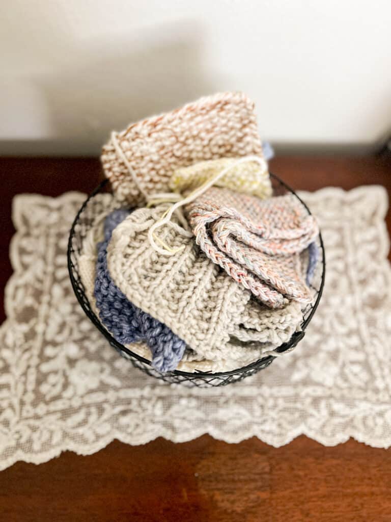 A black wire basket filled with knit swatches sits on top of a dark wooden table with a lace doily.