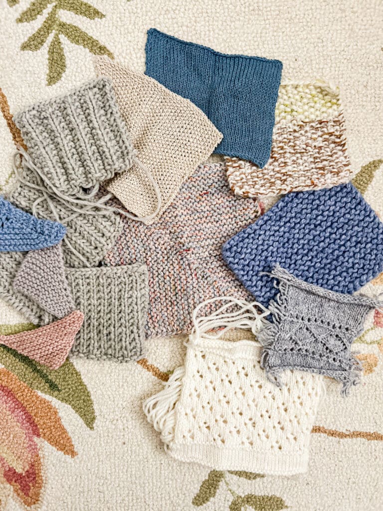 A pile of knit swatches in various pastel colors, some in simple textures and some with lace, some quite large and others very small, sits on top of a cream-colored rug with pink and green floral details on it.