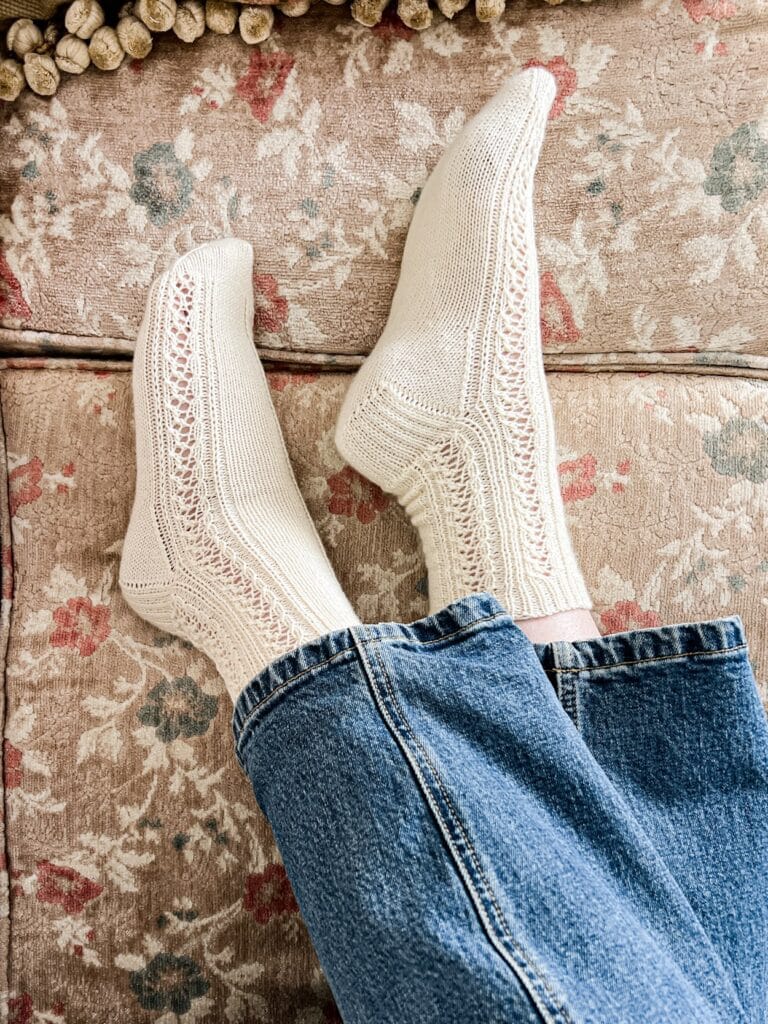 A woman's feet wearing a pair of white knit socks with lace details down the sides rest on a vintage sofa with pastel floral upholstery.