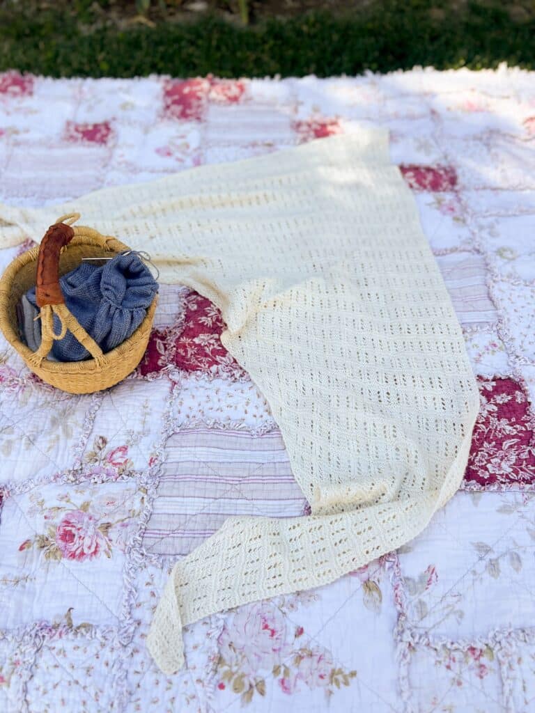A white handknit lacy shawl is spread out on a patchwork quilt in a garden next to a small basket with some blue knitting in progress.