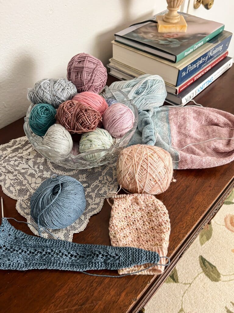 A pile of unfinished knitting projects in various pastel colors sits on a dark wood table next to a cut glass bowl filled with leftover balls of yarn.
