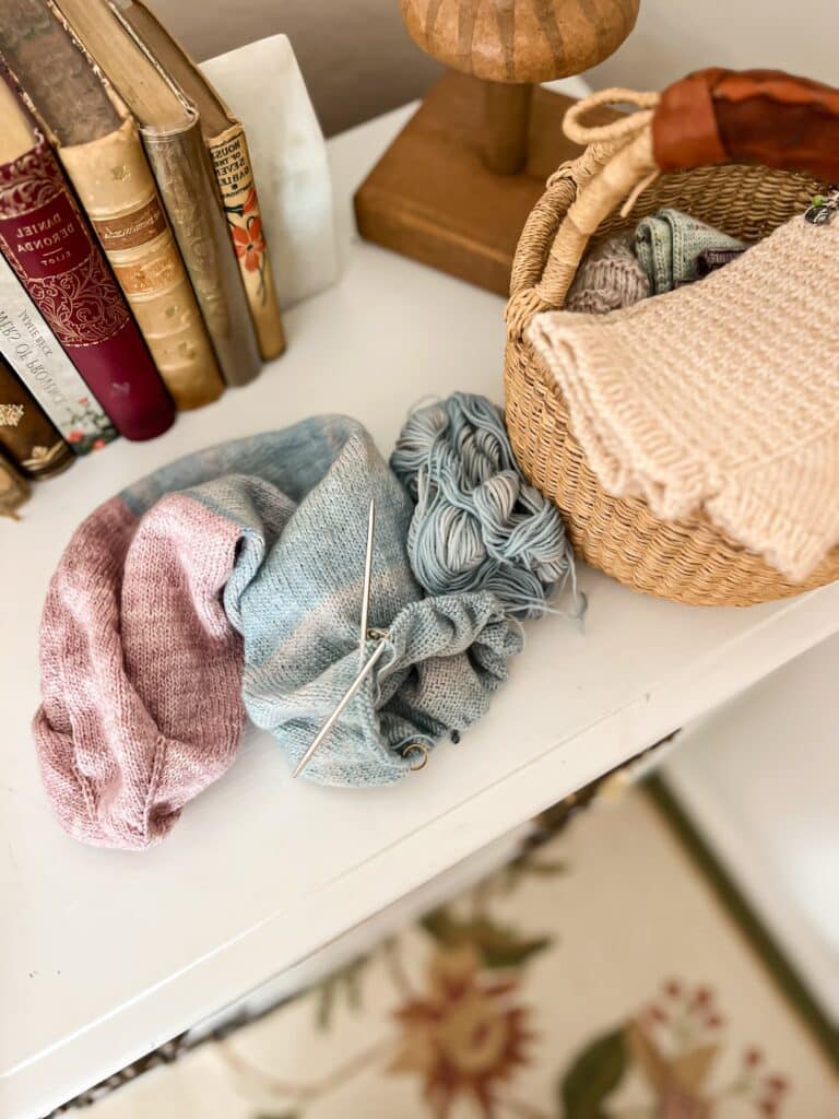 A very long tube of blue and lavender knitting sits on top of a white dresser next to some antique books and a basket with more knitting in it.