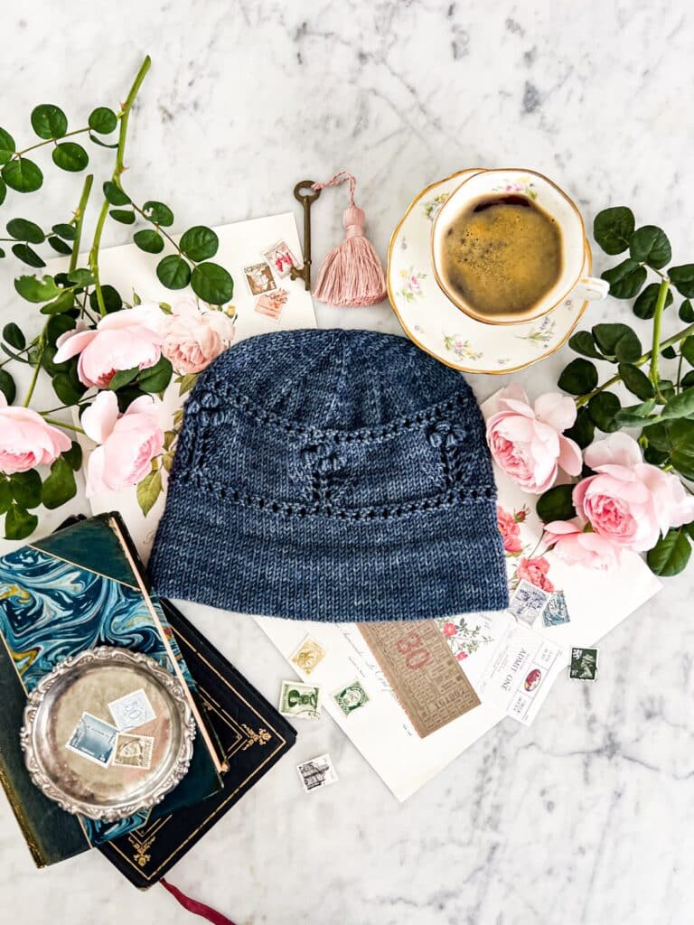 A blue handknit hat with a folded brim and bobbles sits on a white marble countertop surrounded by flowers and books.