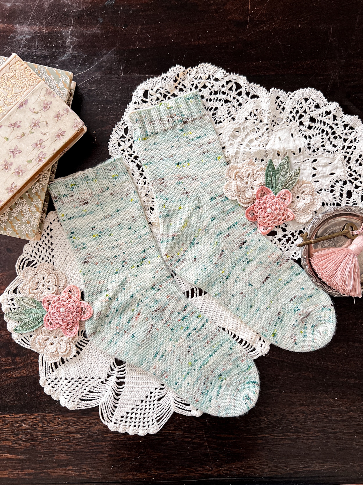 A pair of light green stockinette socks with speckles is laid out on a white doily and a dark wood table. The socks are surrounded by antique books and ribbonwork/crocheted flowers.